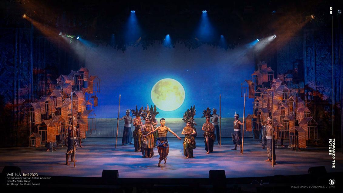 Balinese dancers in headdresses perform under a giant moon, flanked by sculpted stilt houses.