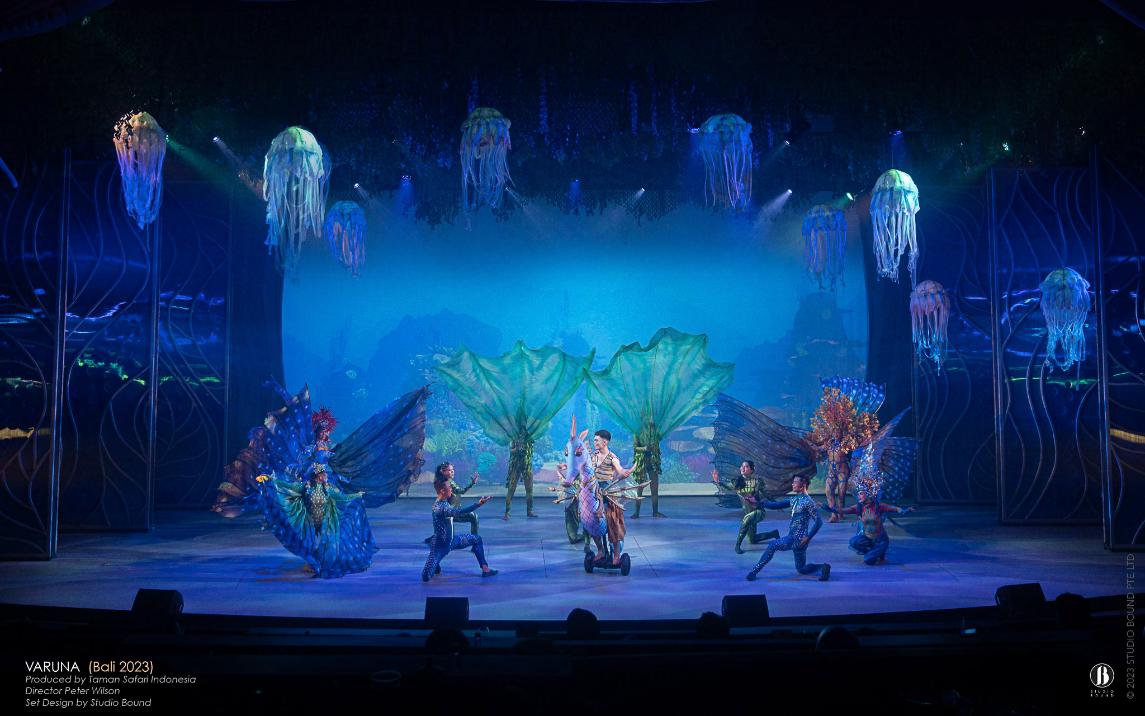 Jellyfish chandeliers and a luminous aquarium wall behind dancers, with a performer riding a seahorse-puppet on stage.