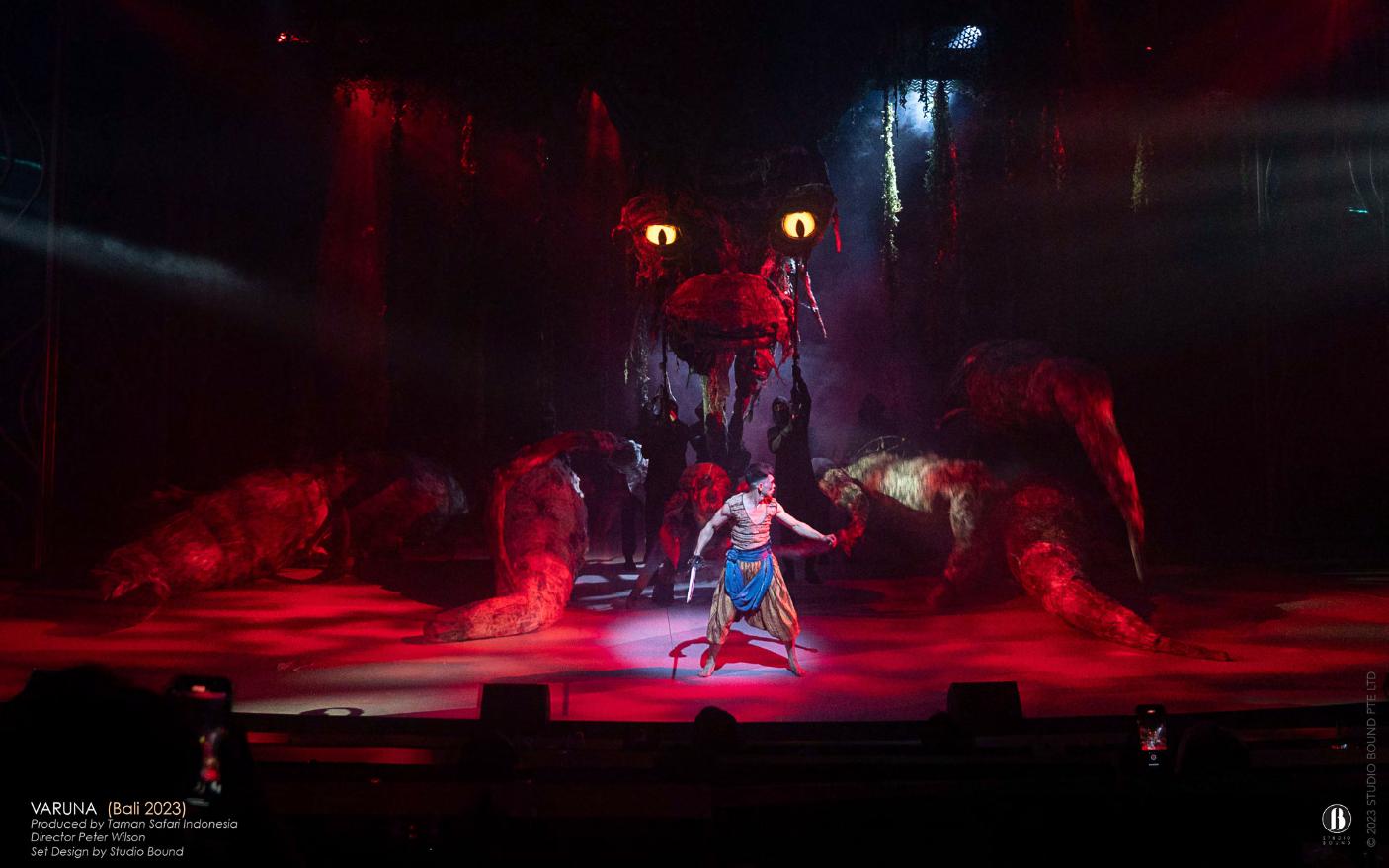 Towering crimson sea-serpent puppet with glowing yellow eyes looming over a lone warrior on stage.