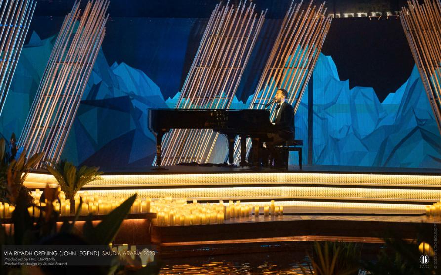 John Legend at a grand piano on a candle-lit stage with a stylised blue mountain landscape projected behind him through bamboo screens.