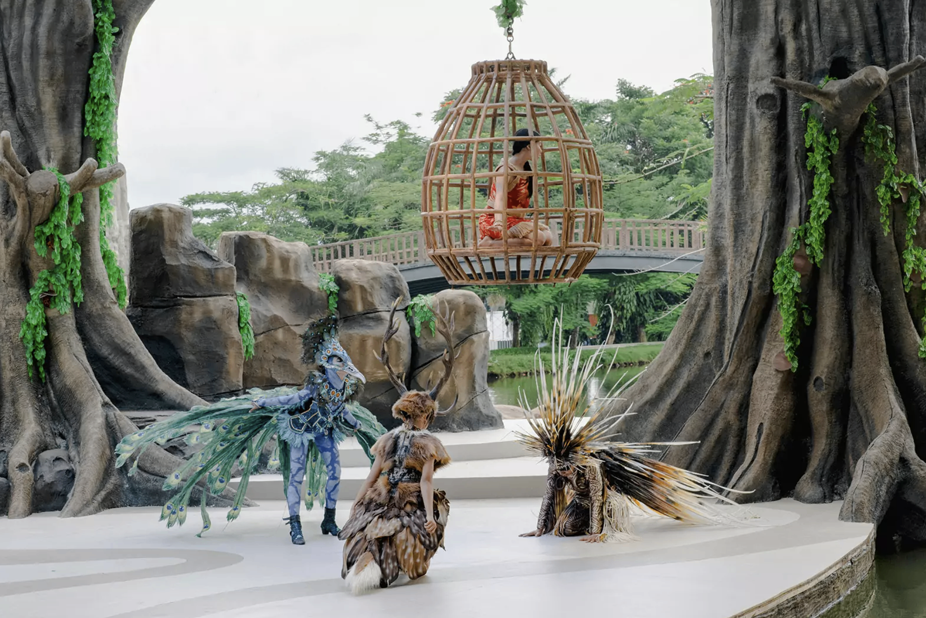 Forest-spirit performers on the Lila stage — a peacock spirit, an antlered creature and a porcupine-quilled figure beneath a suspended rattan cage, framed by sculpted banyan trees.
