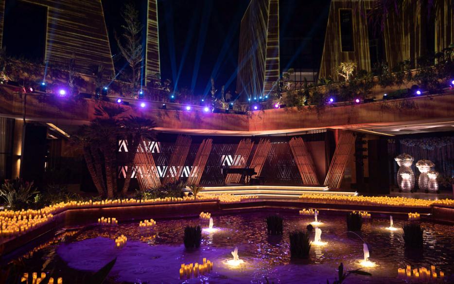 Candle-lit gala stage with bamboo VIA-monogram screen and grand piano, surrounded by pools of water and uplit purple terraces above.