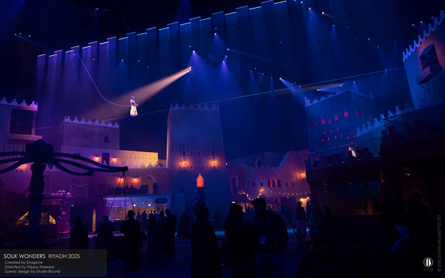 A tightrope walker traverses the city walls under a single shaft of light — the architecture redrawn in cool blue, the design built to perform as cinematically after dark as it does by day. Scenic design by Studio Bound.