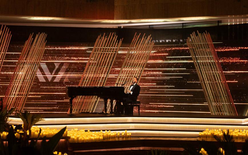 Tight stage view of John Legend at a grand piano with a glowing wall of fire-orange candle light behind tall bamboo screens.