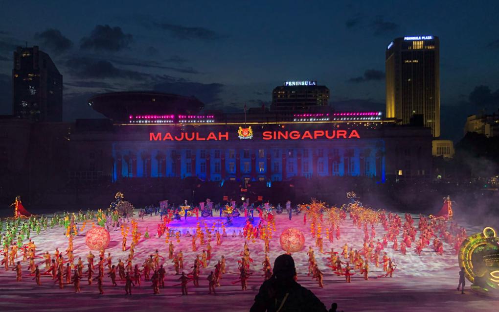 "Majulah Singapura" projected across the Asian Civilisations Museum at dusk, with lantern props and ensemble formations filling the field. Set and prop design by Patrick Larsen, Studio Bound.