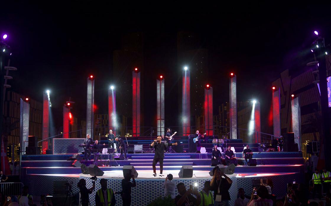 Darb Lusail stage lit in red and cool white, viewed from the crowd with phones raised, performer mid-song on the patterned mosaic apron.