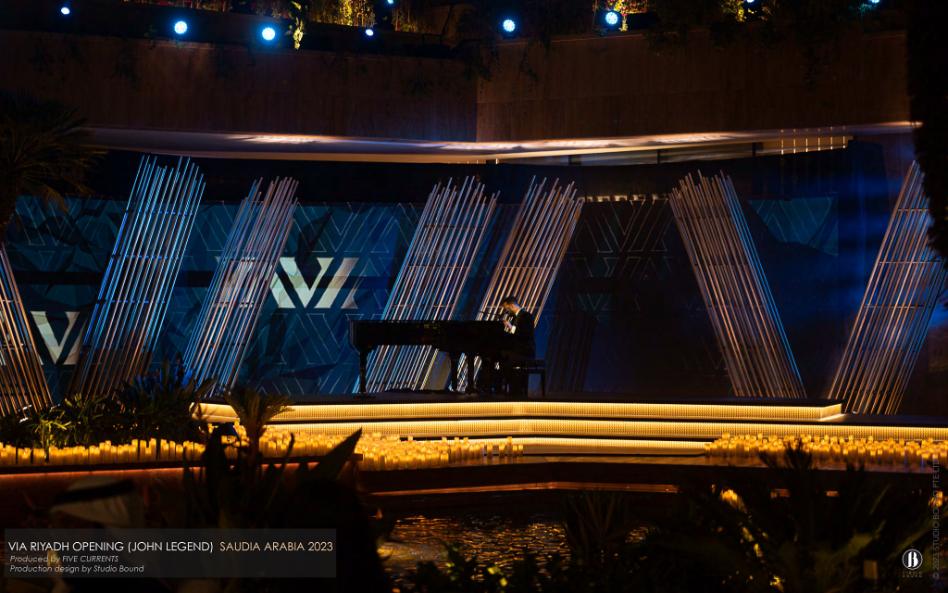 John Legend playing a grand piano on a candle-rimmed stage, framed by an architectural bamboo screen with the VIA Riyadh monogram behind him.