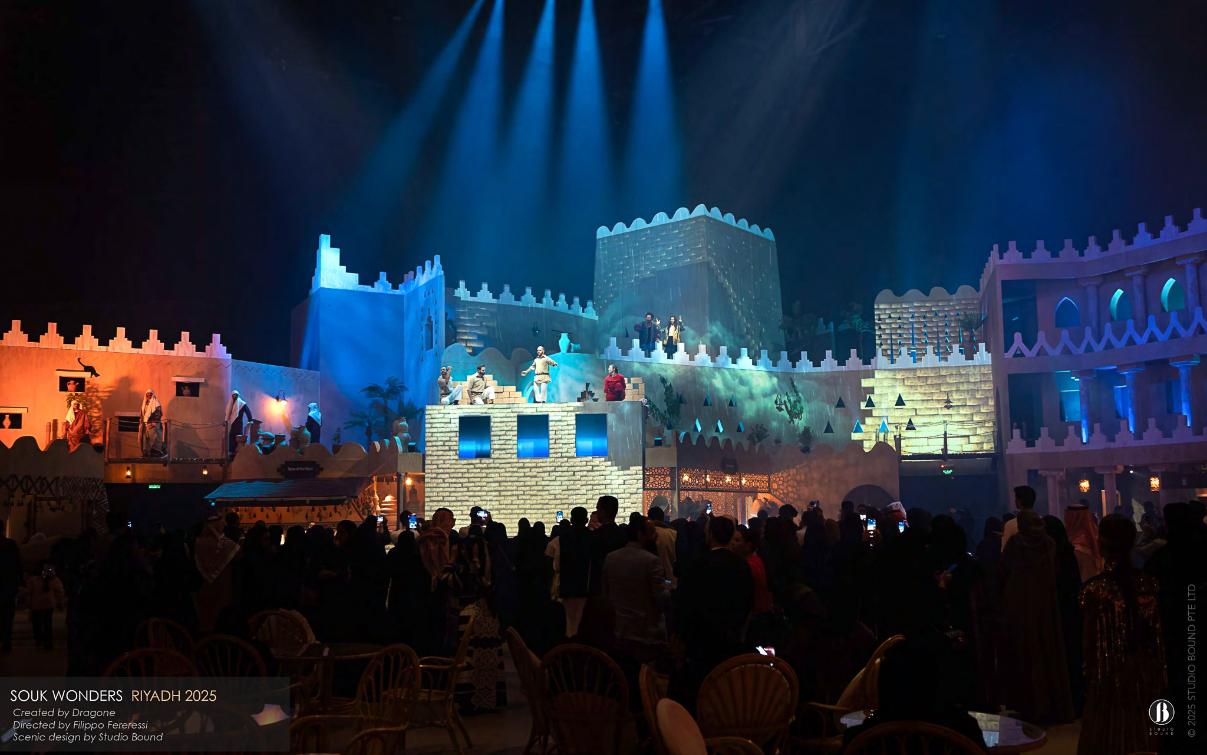 A nighttime set-piece on the upper ramparts — guests gathered close, performers staged across rooftops and battlements, the marketplace itself becoming the auditorium. Scenic design by Studio Bound.