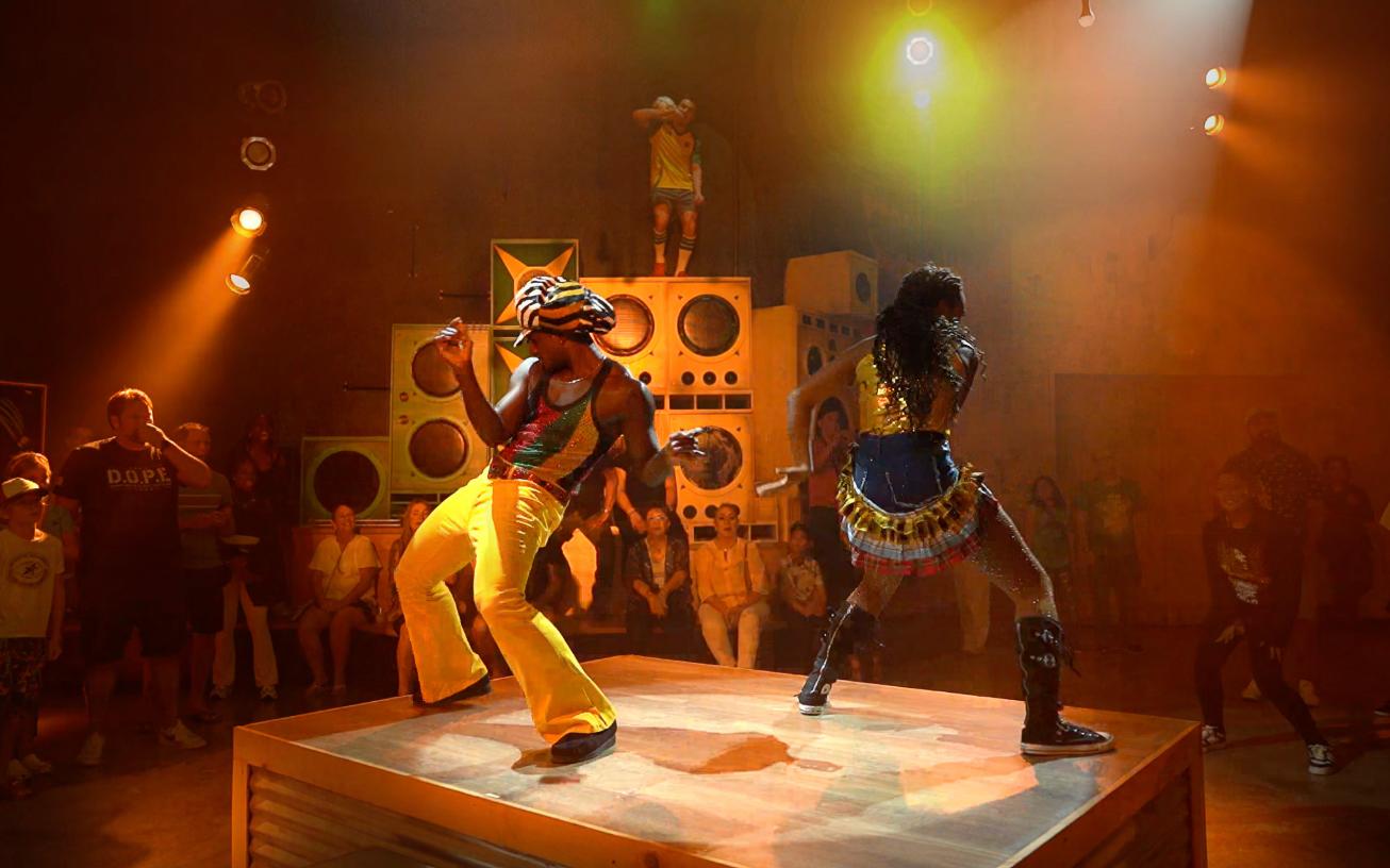 The dancehall — a stacked wall of vintage speakers as set, audience standing inches from the performers. The room designed so the architecture itself is the sound system. Production design by Patrick Larsen, Studio Bound.