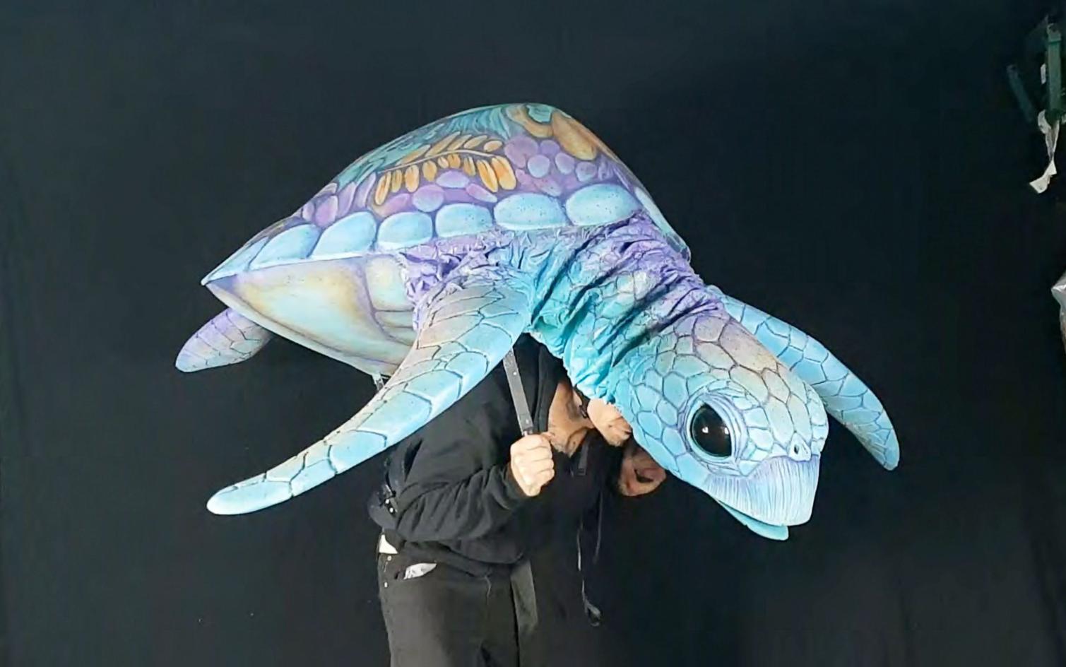 A performer operates a large hand-painted sea turtle puppet in shades of blue and violet against a black backdrop.