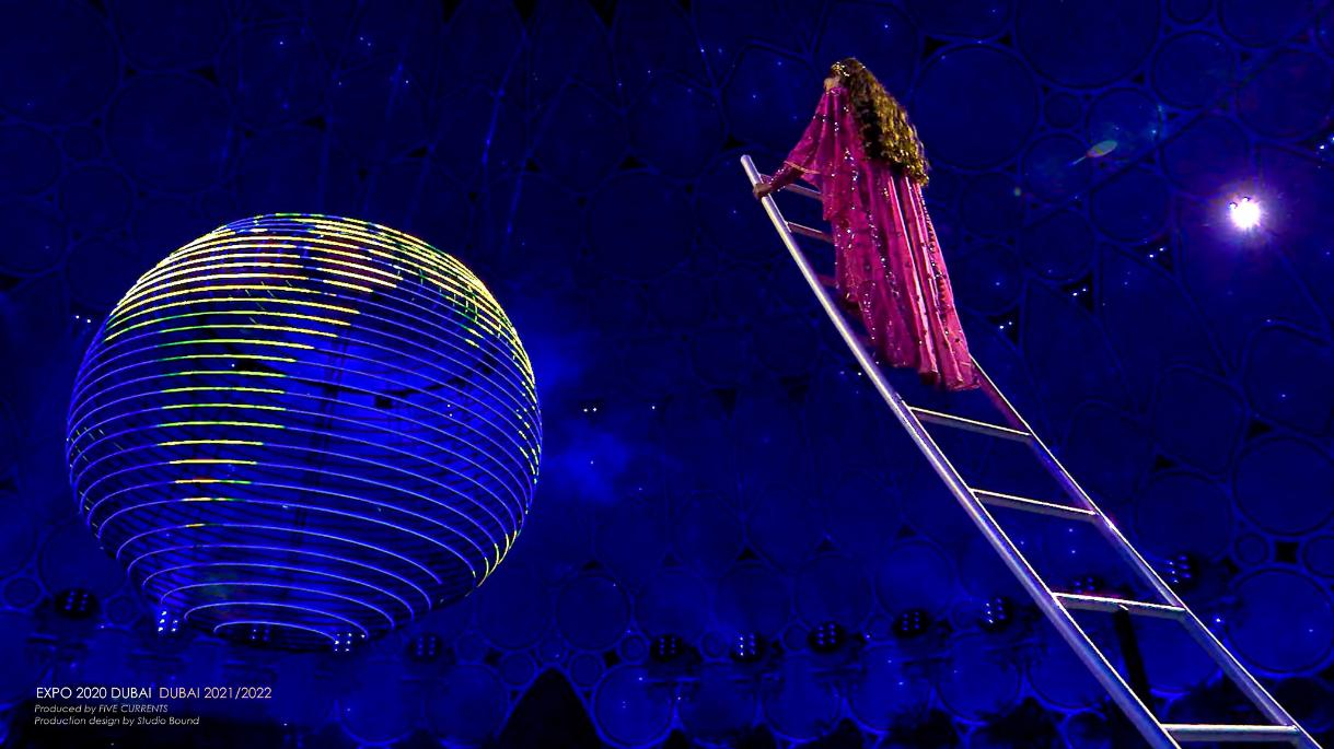 A performer climbs a ladder toward a luminous sphere suspended inside Al Wasl — a single figure used to scale a 67-metre-high room. Production design by Studio Bound.