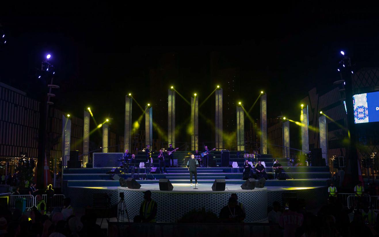 Darb Lusail boulevard stage washed entirely in golden yellow, with sharp top-light beams over the band and crowd in silhouette.