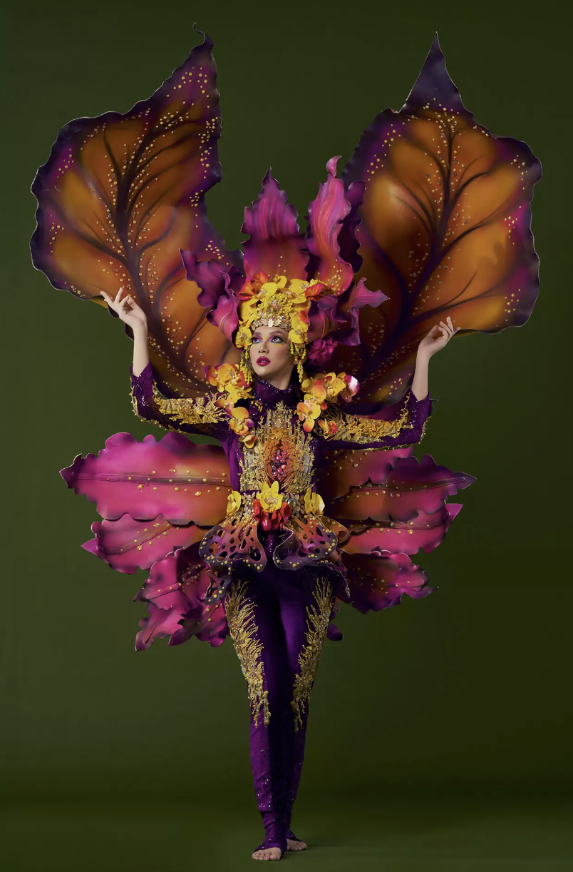 Lila performer in an orchid-and-butterfly costume with sculpted amber wings, magenta petal skirt and a floral golden headpiece against a green backdrop.