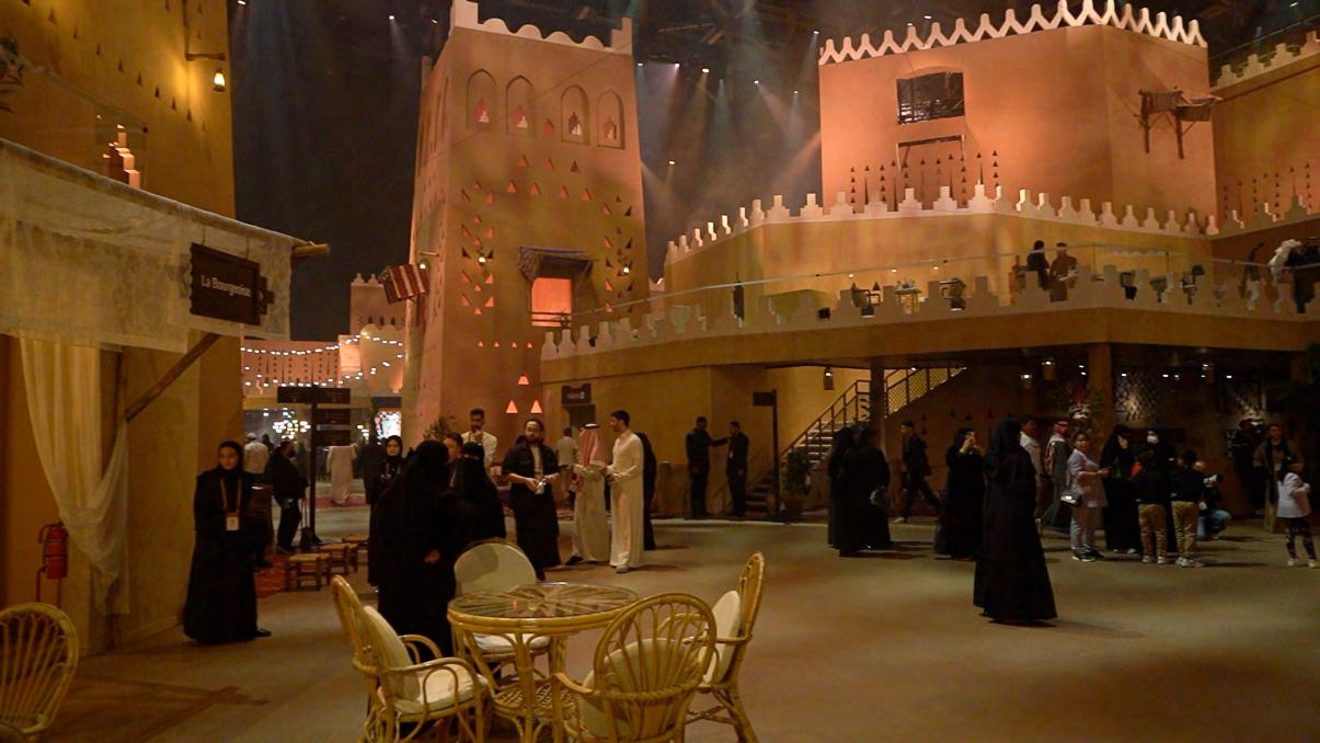Street level inside the souk — La Bourgade quarter, with guests moving between mudbrick façades, lanterns and rattan café settings. The labyrinth working as a place to linger, not just to pass through. Production design by Patrick Larsen, Studio Bound.