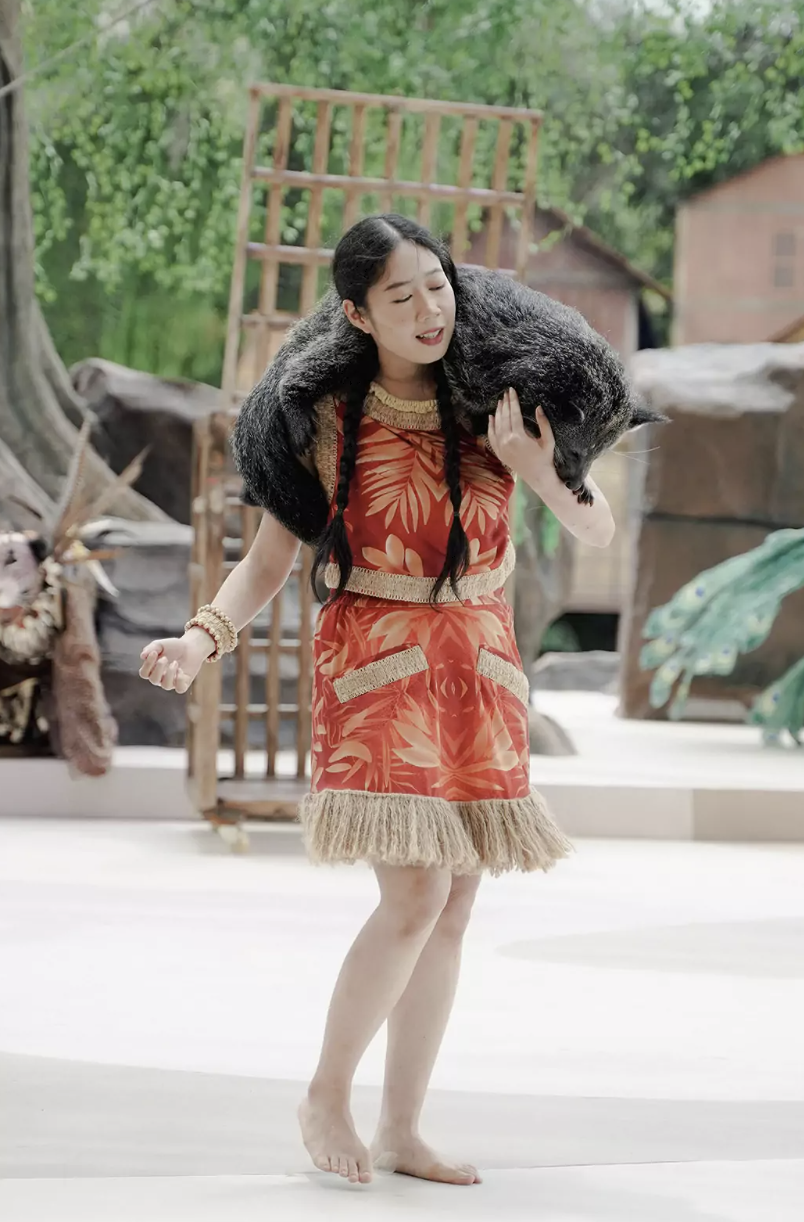 Lila on stage in a red palm-print dress with woven trim and fringe, carrying a binturong on her shoulders against the forest set.