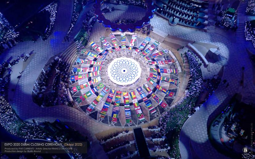 Aerial of the Closing Ceremony — a ring of national flags arranged on the plaza floor, the ceremony composed for the camera looking straight down as much as for the eye looking up. Production design by Studio Bound.