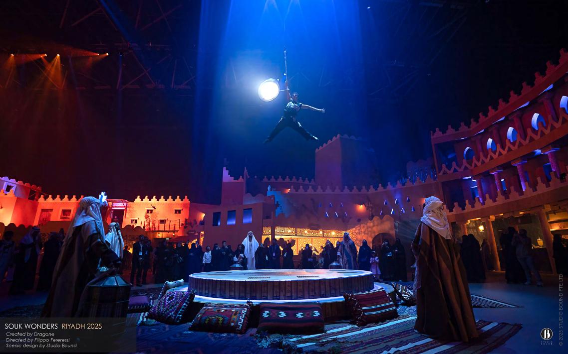An aerialist crosses the souk on a luminous drum, lit against a wash of deep blue — vertical theatre layered above the horizontal life of the market. Scenic design by Studio Bound.