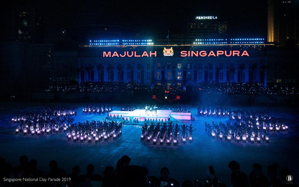 The same façade after dark — projection-lit drummers and a central plinth stage hold the field below "Majulah Singapura." Set and prop design by Patrick Larsen, Studio Bound.