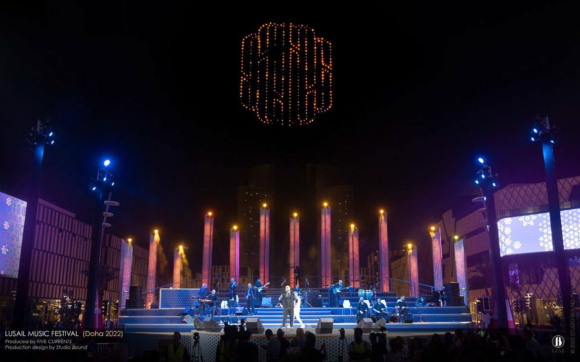 Night view of the Darb Lusail stage in warm amber and rose, with an Arabic calligraphy drone formation glowing above the towers.