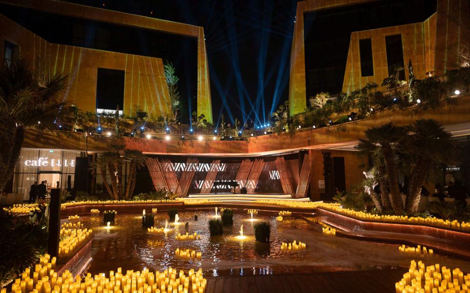 Sunken candle-lit gala stage seen across reflecting pools and palm trees, framed by warmly-lit limestone facades and skybeams.