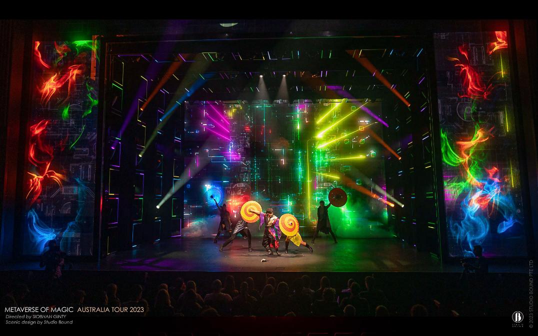 A full-colour ensemble number — fan-and-umbrella choreography lit against side panels of red, green and blue smoke, the digital surround at its brightest. Production design by Patrick Larsen, Studio Bound.