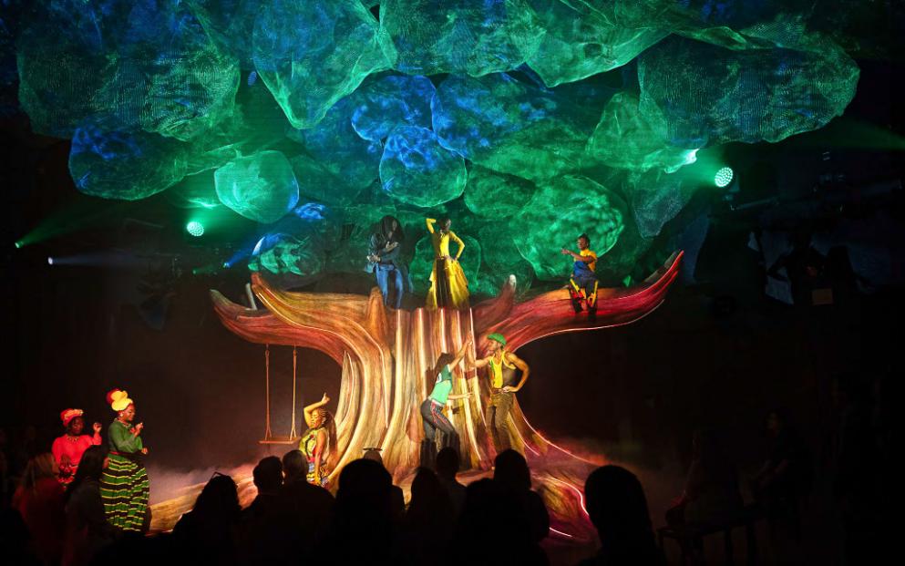 The Jammin' Tree — a sculpted trunk rising out of the floor under a vast canopy of green and blue mesh "leaves," performers staged in its branches. The room that turns the audience into a circle around a single living object. Scenic design by Studio Bound.