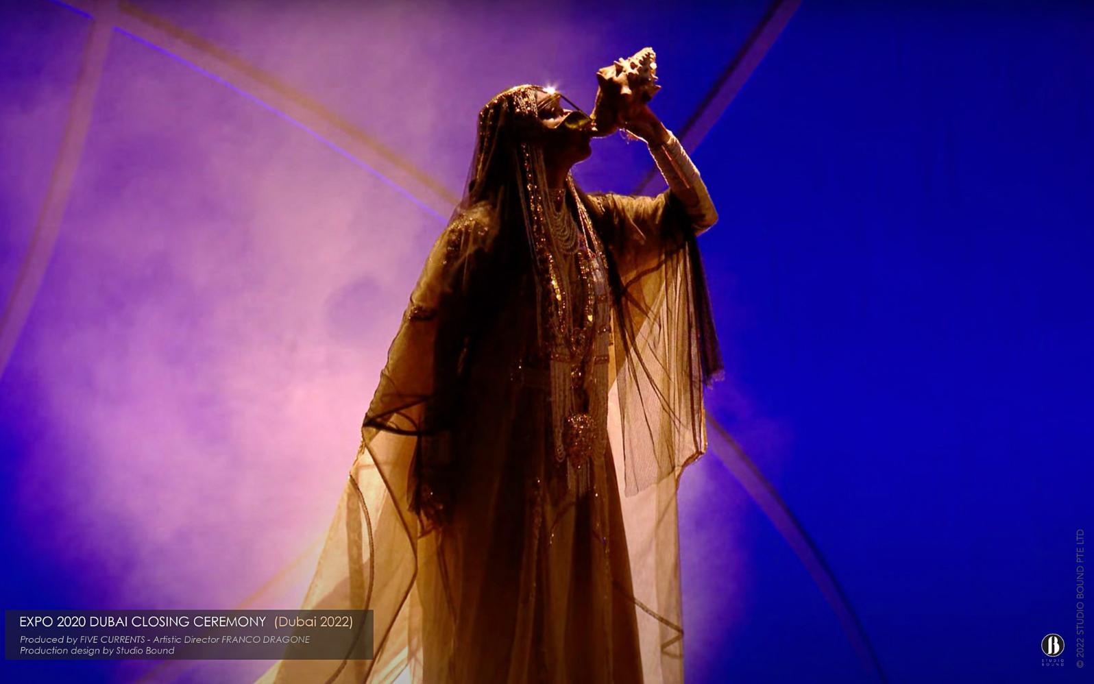 The closing solo — a single veiled performer with a conch shell against a wash of violet light. The whole 130-metre room, in the end, focused down to one figure. Production design by Studio Bound.