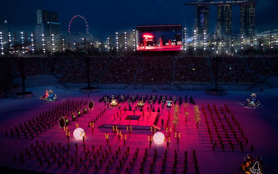 Marina Bay backdrop — the Singapore Flyer and Marina Bay Sands frame the field as performers, lit floats and lantern props fill The Padang in formation. Set and prop design by Patrick Larsen, Studio Bound.