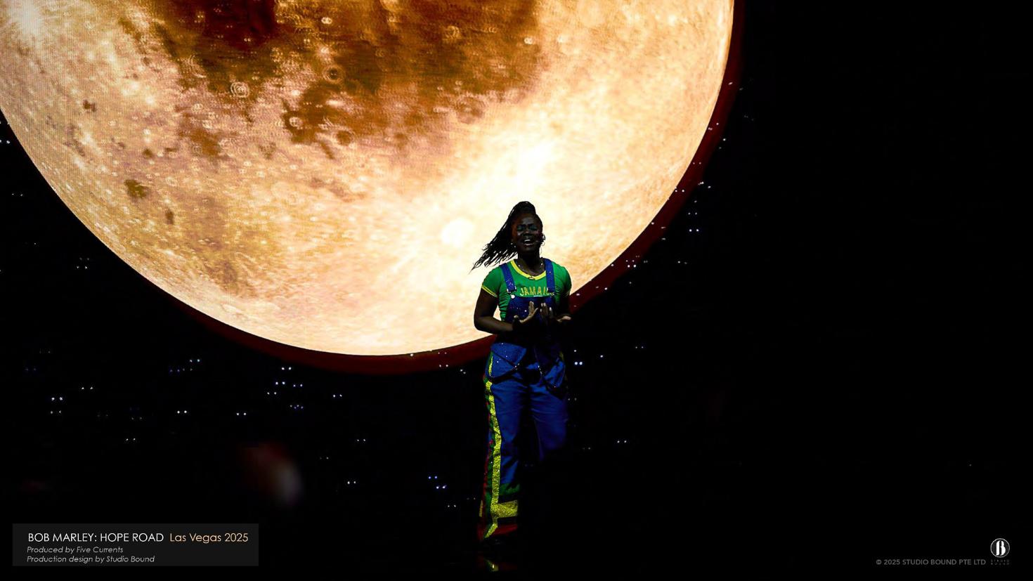 The finale — a single performer in Jamaican colours silhouetted against a vast rising moon, the journey resolving from many rooms into one image. Production design by Patrick Larsen, Studio Bound.