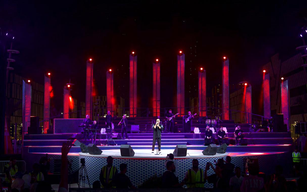 Darb Lusail boulevard stage in deep red, with a soloist at the centre microphone backed by a full band on tiered steps.