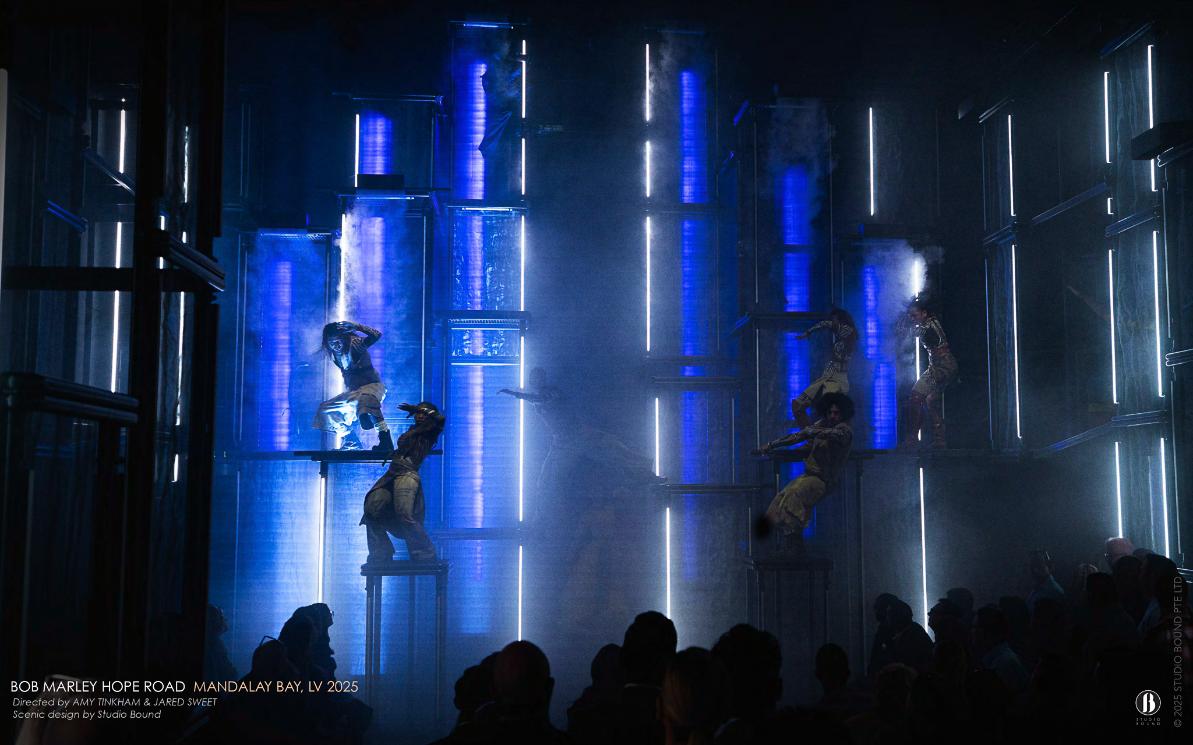 The cathedral of light in blue — kinetic light columns and atmospheric haze turning a room into weather, performers climbing the architecture rather than standing in front of it. Scenic design by Studio Bound.