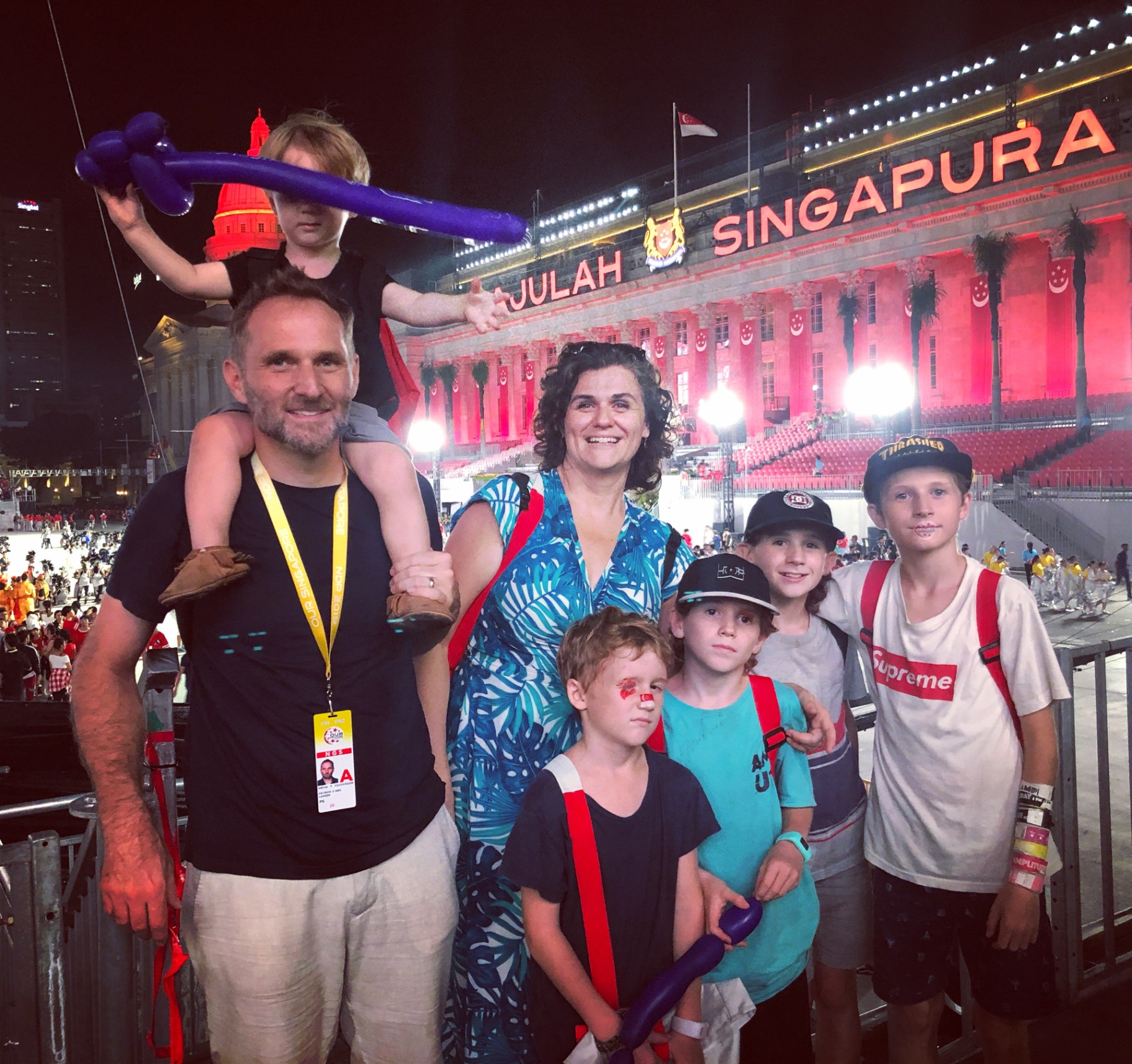 Patrick Larsen with family at a Singapore National Day event under illuminated "SINGAPURA" signage.