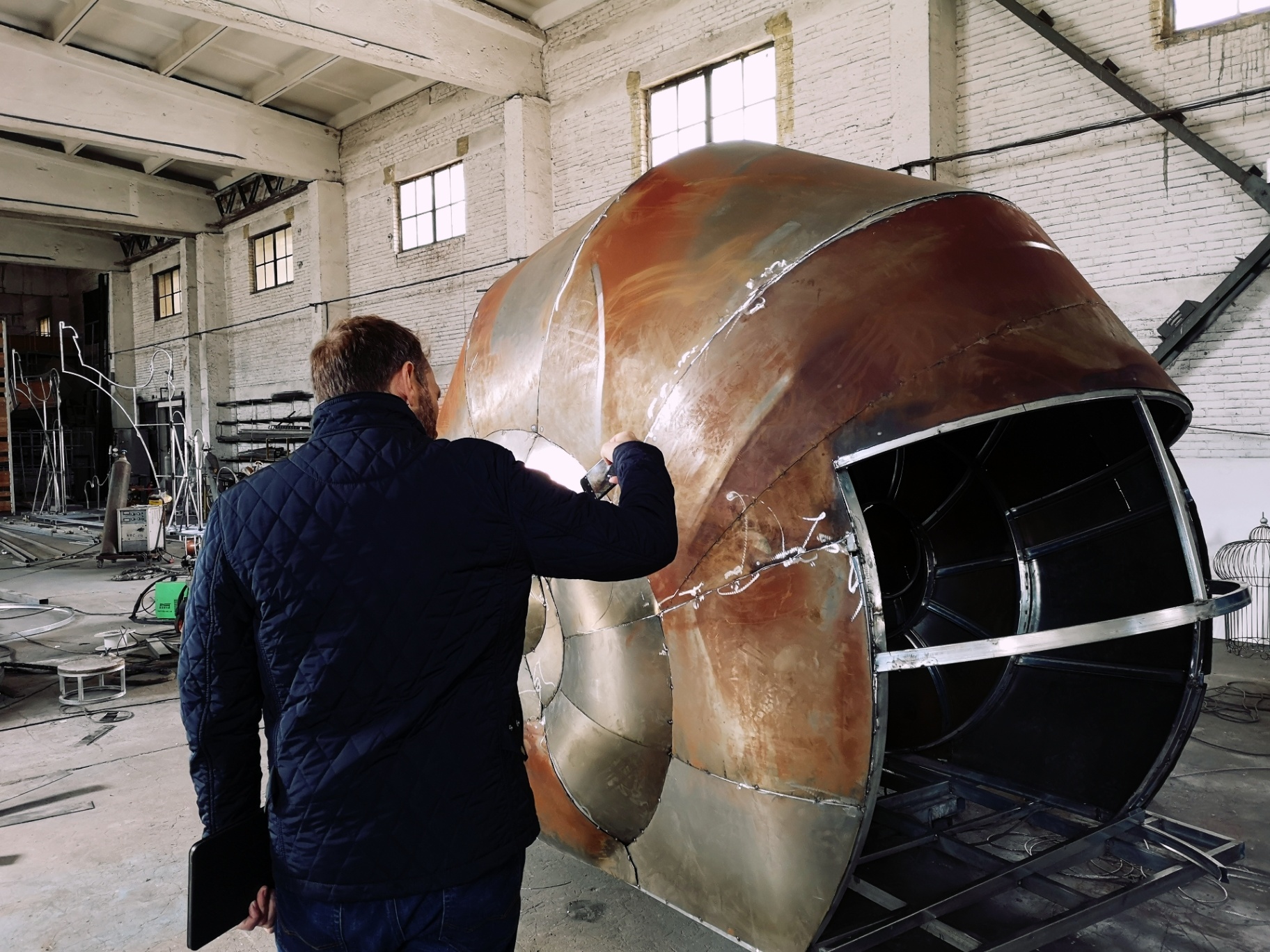Patrick Larsen inspects a large curved metal shell structure in a fabrication workshop.